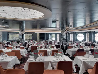 Das Golden Sand Restaurant an Bord der MSC Seaview. Viele weiss gedeckte Tische mit braunen Stühlen und Weingläsern. Grosse Bullaugen mit Blick auf das Wasser.