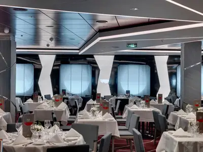 Elegantes Silver Dolphin Restaurant der MSC Seaview. Viele weiß gedeckte Tische, graue Stühle und Weingläser. Stilvolle Deckenbeleuchtung und X-förmige Wandelemente. Grünes Exit-Schild.