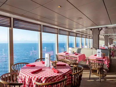 Ein Innenbereich des Ocean Point Restaurants an Bord der MSC Seaview mit Tischen und Stuehlen. Rote, karierte Tischdecken und grossen Fenstern bieten eine weite Aussicht auf das Meer und den Horizont.