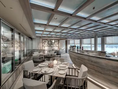 Ein elegantes Restaurant an Bord der MSC Seaview mit stilvoller Einrichtung, maritimem Kunstwerk und Meerblick auf Yachten. Gedeckte Tische und eine Bar.