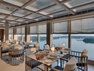 Ein helles und elegantes Restaurant an Bord der MSC Seaview mit modernen Tischen, bequemen Stühlen und großen Fenstern. Die Tische sind gedeckt und mit dekorativen Objekten verziert. Im Hintergrund ist ein ruhiger Yachthafen mit Booten und grünen Ufern zu sehen.