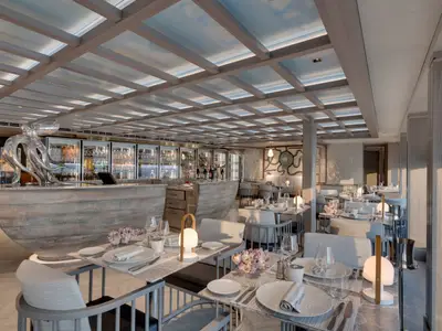 Ein elegantes, helles Restaurant an Bord der MSC Seaview. Im Vordergrund sind Tische mit weißen Gedecken und modernen Tischlampen zu sehen. Links steht eine auffällige, silberne Oktopus-Skulptur auf einer geschwungenen Bar. Im Hintergrund sind gekühlte Glasvitrinen mit Getränken und ein Wandgemälde in Form eines Oktopus zu erkennen. Das Deckengitter lässt diffuses Licht durch.