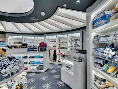 Heller, moderner MSC Shop an Bord der MSC Seaview. Weisse Regale zeigen gefaltete Kleidung, Taschen, Plueschtieren (Teddybaeren, Maskottchen) und Souvenirs. Eine geschwungene Decke und gemusterter Teppichboden.