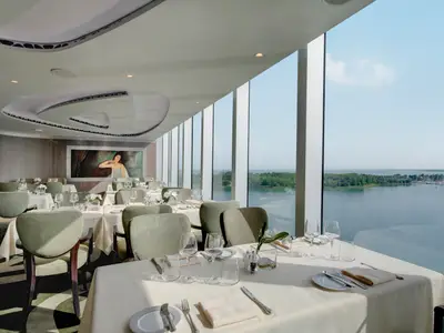 Elegantes Restaurant an Bord der MSC Seaview mit weissgedeckten Tischen, olivgrünen Stühlen, Wandgemälde. Grosse Fenster zeigen Meer, eine bewaldete Insel und Boote.