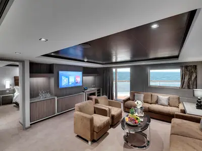 Eine luxuriöse Suite an Bord der MSC Seaview. Der moderne Wohnbereich mit braunen Sofas, TV und frischem Obst bietet Meerblick. Links ist ein Schlafzimmer sichtbar.