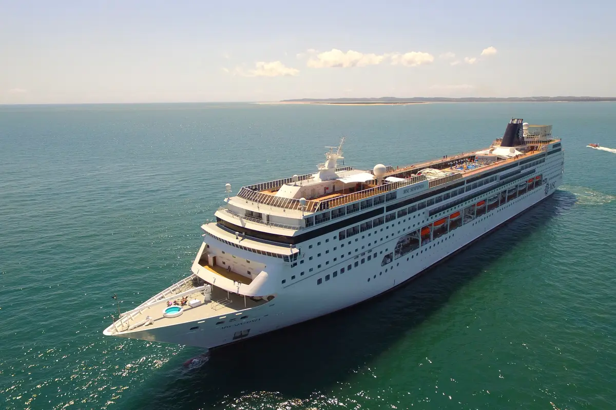 Vogelperspektive der MSC Sinfonia, die auf blaugrünem Meer unter einem klaren Himmel fährt. Ein Beiboot folgt ihr hinten rechts, während im Hintergrund eine sandige Inselkette sichtbar ist.