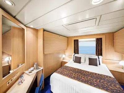 Eine helle Junior Aussenkabine an Bord der MSC Sinfonia mit einem Doppelbett, einem Schreibtisch mit Spiegel, zwei Beistelltischen und einem Fenster mit Blick auf das Meer. Die Einrichtung ist in hellem Holz gehalten.