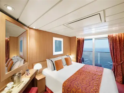 Eine gemütliche Balkonkabine an Bord der MSC Sinfonia mit einem ordentlich gemachten Doppelbett, Nachttischen und einem Schreibtisch mit Spiegel. Durch die Balkontür ist das Meer sichtbar. Braune Vorhänge hängen seitlich.