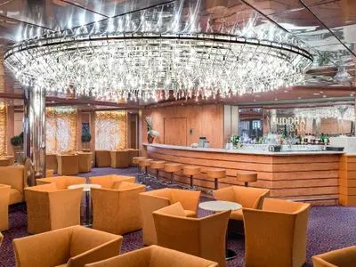 Blick in die elegante Buddha Bar an Bord der MSC Sinfonia. Ein riesiger Kronleuchter hängt über einem Tresen mit vielen Getränken und orangefarbenen Lounge-Sesseln.
