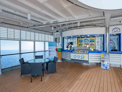 Eine helle Eisdiele namens Sammontana an Bord der MSC Sinfonia mit blauer Theke, bunten Eissorten und Tischen. Grosse Fenster bieten einen weiten Blick auf das offene Meer.