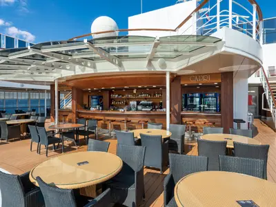 Die Capri Bar an Bord der MSC Sinfonia: Holztresen mit Getränken, Barhockern, Rattan-Tischen und Stühlen auf dem Holzdeck. Glasdach über der Bar, im Hintergrund das Meer.