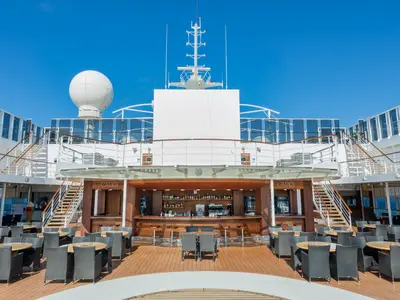Blick auf die Capri Bar an Bord der MSC Sinfonia. Eine holzverkleidete Aussenbar mit Barhockern. Davor Tische und Stühle auf Holzdeck. Im Hintergrund obere Decks unter blauem Himmel.