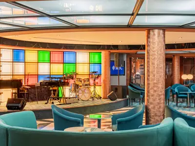 Die bunte Manhattan Bar an Bord der MSC Sinfonia zeigt eine Bühne mit Klavier und Schlagzeug. Helle Stühle um kleine Tische laden zum Verweilen ein.