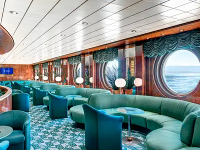 Manhattan Bar an Bord der MSC Sinfonia mit gruenen, runden Sofas, Sesseln und kleinen Tischen, umgeben von Holzvertäfelung und grossen Bullaugen, die den Meerblick zeigen. Eine gemuetliche Atmosphaere.