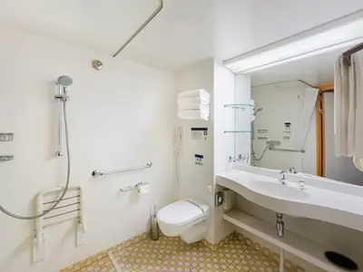 Barrierefreies Badezimmer an Bord der MSC Sinfonia. Es zeigt eine Dusche mit Klappsitz und Haltegriffen, Toilette, Waschbecken und Spiegel. Helle, moderne Einrichtung mit Chromarmaturen.