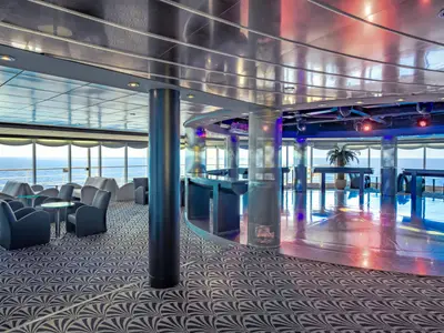Innenansicht der Pasha Club Disco an Bord der MSC Sinfonia. Eine Lounge mit Sesseln und Tischen, dahinter eine Bar und Tanzfläche mit reflektierendem Boden. Große Fenster bieten weiten Meerblick.