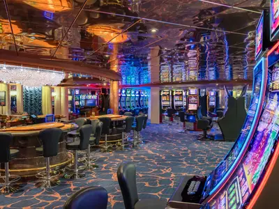 Sanremo Casino an Bord der MSC Sinfonia. Viele bunte Spielautomaten, Spieltische und ein großer Kristall-Kronleuchter. Die spiegelnde Decke reflektiert lebhafte Lichter.