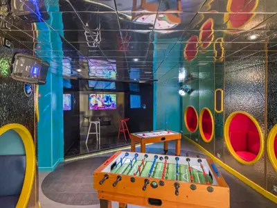 Spielraum an Bord der MSC Sinfonia mit Tischkicker, Air-Hockey und runden, farbigen Sitznischen. Großer Bildschirm und spiegelnde Decke. Moderne und lebhafte Atmosphäre.