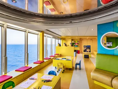Heller LEGO-Junior-Club an Bord der MSC Sinfonia mit großen Fenstern zum Meer. Gelbe Tische, Bänke mit roten Kissen, grüne Sofas und LEGO-Dekorationen sind sichtbar.
