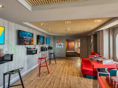 Innenansicht des Teen Clubs an Bord der MSC Sinfonia mit mehreren Bildschirmen für Videospiele, bequemen roten Sofas und Hockern sowie einem Meerblick durch das ovale Fenster. Die Wände sind hellgrau holzvertäfelt und mit Neon-Schriftzügen verziert.