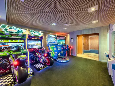 Geräumige Spiele-Arkade an Bord der MSC Sinfonia mit mehreren Arcade-Automaten, darunter zwei Motorrad-Rennsimulatoren, ein Snowboard-Simulator und ein Greifautomat. Die Automaten sind hell beleuchtet.