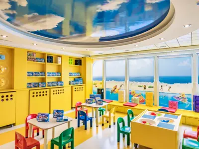 Ein farbenfroher Lego-Themen-Spielbereich an Bord der MSC Sinfonia mit gelben Möbeln, Tischen, Stühlen und einem Spieltisch. Große Fenster bieten einen Blick auf das Meer und sind mit Lego-Szenen dekoriert.