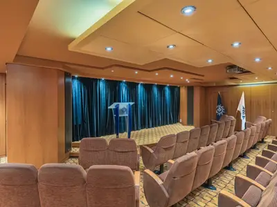 Innenansicht eines Konferenzraums an Bord der MSC Sinfonia mit Reihen brauner Stoffsitze. Vorne eine Bühne mit Rednerpult, dahinter blaue Vorhänge. Zwei Fahnen stehen daneben.