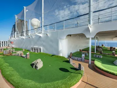 Ein Minigolfplatz im Freien an Bord der MSC Sinfonia. Grüne Kunstrasenbahnen mit Felsen und Holzstämmen als Hindernisse. Im Hintergrund die weisse Bordwand des Schiffes und das Meer.