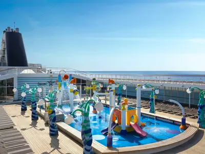 Der farbenfrohe Doremi Spray Park an Bord der MSC Sinfonia auf dem Sonnendeck. Ein Wasserspielplatz mit Rutschen, Fontänen und bunten Figuren. Im Hintergrund blauer Himmel, das weite Meer und Sonnenliegen.
