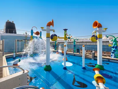 Bunter Doremi Spray Park auf der MSC Sinfonia mit spritzenden Wasserspielgeräten und flachem Becken. Liegestühle und Schornstein im Hintergrund unter blauem Himmel.