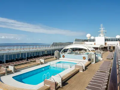 Das sonnige Pooldeck der MSC Sinfonia mit zwei tuerkisfarbenen Pools und vielen Liegestuehlen. Im Hintergrund ist das Meer und eine Kuestenlinie unter blauem Himmel sichtbar.