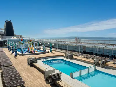 Das sonnige Oberdeck der MSC Sinfonia mit einem großen Pool und farbenfrohem Doremi Spray Park für Kinder. Viele Liegestühle auf dem Holzdeck. Im Hintergrund das Meer und eine Küste unter blauem Himmel.