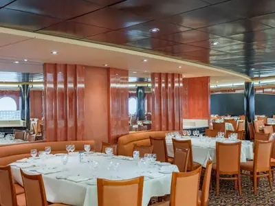Das Restaurant Il Galeone an Bord der MSC Sinfonia zeigt elegant gedeckte Tische, orangefarbene Stühle und Sitzbänke. Spiegel und ein Bullauge sind an den Wänden sichtbar.