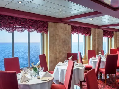 Blick in das elegante Restaurant Il Covo an Bord der MSC Sinfonia. Mehrere gedeckte Tische mit roten Stuehlen stehen vor grossen Fenstern, die einen weiten Blick auf das offene Meer bieten. Der Raum ist in Rottönen gehalten.