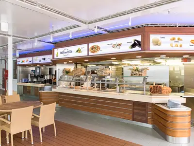 Das helle Buffetrestaurant La Terrazza an Bord der MSC Sinfonia. Langer Tresen mit Speisen und Getränken. Moderne Tische und Korbstühle. Beleuchtete Info-Tafeln für Gäste.