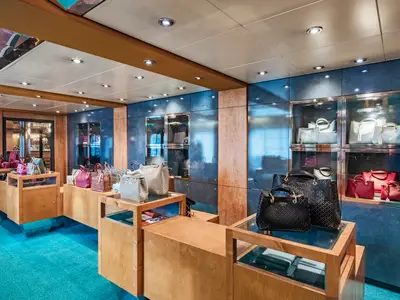 Shop an Bord der MSC Sinfonia. Elegante Handtaschen auf Holztheken und in Glasvitrinen. Blauer Teppich, Wände aus Holz und blauen Paneelen, helles Ambiente.