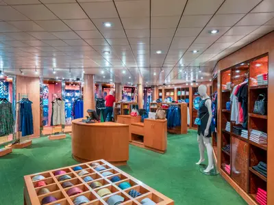 Helle Boutique an Bord der MSC Sinfonia mit grünem Teppich und Holzeinrichtung. Mannequins, Kleiderständer und Auslagen zeigen Kleidung, Taschen und Krawatten.