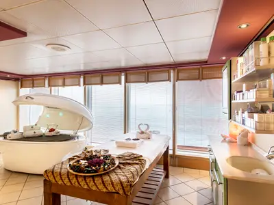 Heller Spa-Behandlungsraum an Bord der MSC Sinfonia mit Hydrotherapiebett und Massageliege. Blick auf das Meer durch Fenster, rechts Produkte und Waschbecken.