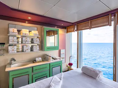 Ein heller Spa-Behandlungsraum an Bord der MSC Sinfonia mit einer Liege, einem Waschbecken und grossen Fenstern mit Blick auf das weite Meer. Gestapelte Handtücher mit 'Aurea Spa' Logo sind sichtbar.