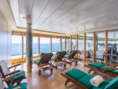 Entspannungsbereich an Bord der MSC Sinfonia. Holzliegen mit grünen Polstern bieten weiten Meerblick. Durch eine Glaswand ist ein Fitnessraum mit Sportgeräten sichtbar.