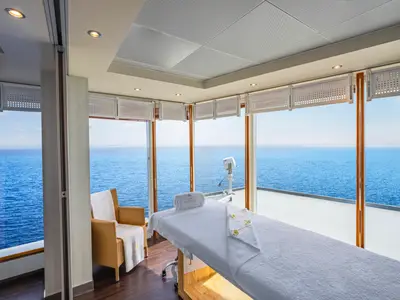 Ein heller Massageraum im MSC Aurea Spa an Bord der MSC Sinfonia mit einer Massagebank, einem Sessel und großen Fenstern, die einen weiten Blick auf das offene Meer bieten.