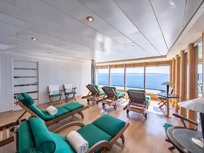 Ein heller Entspannungsraum im MSC Aurea Spa der MSC Sinfonia mit mehreren Holzliegen und grünen Auflagen. Große Fenster bieten einen weiten Blick auf das offene Meer und den Horizont. Einige kleine Tische und Stühle sind ebenfalls sichtbar.