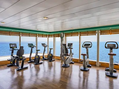 Fitnessstudio an Bord der MSC Sinfonia mit Technogym-Heimtrainern und weitem Meerblick durch große Fenster. Holzboden, helle Decke. Kein Personal oder Passagiere zu sehen.