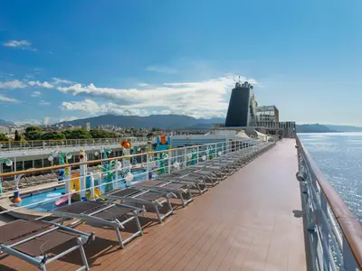 Sonnendeck der MSC Sinfonia. Links bunter Kinder-Wasserpark, viele leere Liegen. Rechts ein Gehweg mit Meerblick. Dahinter Küstenstadt, Berge und blauer Himmel.