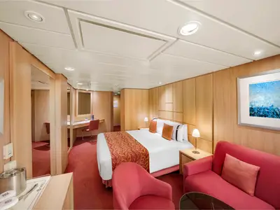 Eine gemuetliche Junior Suite Aurea an Bord der MSC Sinfonia mit Doppelbett, rotem Teppich, Sofa und Holzeinrichtung. Die Kabine hat eine Schminkecke und eine helle, einladende Atmosphaere.