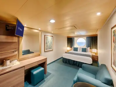 Gemütliche Aussenkabine an Bord der MSC Splendida mit Doppelbett, blauen Kissen und einem Porthole mit Meerblick. Links sind Schminktisch, Spiegel und ein Wandfernseher zu sehen.