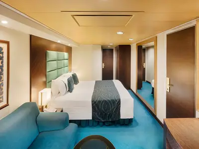 Eine moderne Balkonkabine an Bord der MSC Splendida, ausgestattet mit einem bequemen Doppelbett, einem blauen Sofa und einer hellen, freundlichen Einrichtung. Ein Spiegel reflektiert den Eingangsbereich der Kabine.