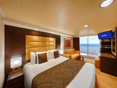Eine helle Premium-Balkonkabine der MSC Splendida mit großem Doppelbett, brauner Tagesdecke und goldener Polsterung, gemütlichem Sofa und Balkon mit Meerblick. Ein Fernseher zeigt das MSC-Logo.