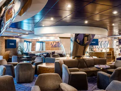 Eine elegante Bar-Lounge an Bord der MSC Splendida mit bequemen Sesseln, Sofas und einem markanten Rezeptionsbereich. Portholes und Bildschirme mit dem MSC Logo schmücken den Raum.