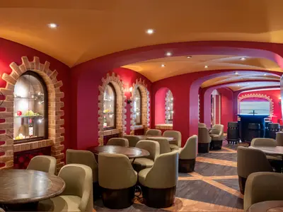 Blick in die rustikal gestaltete L'Enoteca Weinbar an Bord der MSC Splendida, mit roten Wänden, goldenen Deckenwölbungen, zahlreichen Tischen und bequemen, hellbraunen Stühlen. Dekorative Backsteinbögen rahmen Vitrinen mit Weinflaschen und Obst ein. Im Hintergrund eine Bar mit fassähnlichen Hockern.